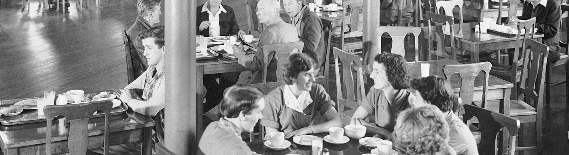 A black and white image of several groups of people sitting in chairs at tables, eating and drinking. The clothing and hairstyles are old fashioned.
