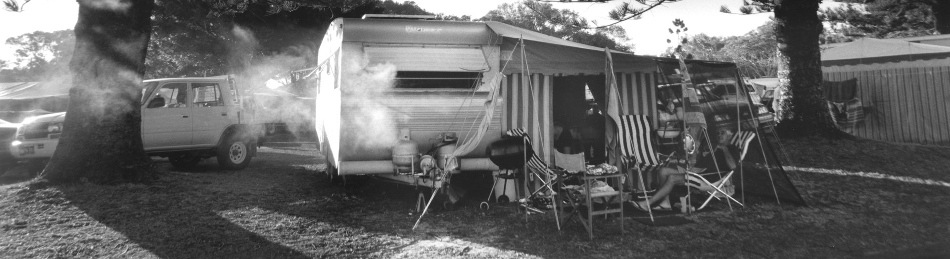 A black and white image of a caravan with an awning and some camp chairs, one occupied by an older man