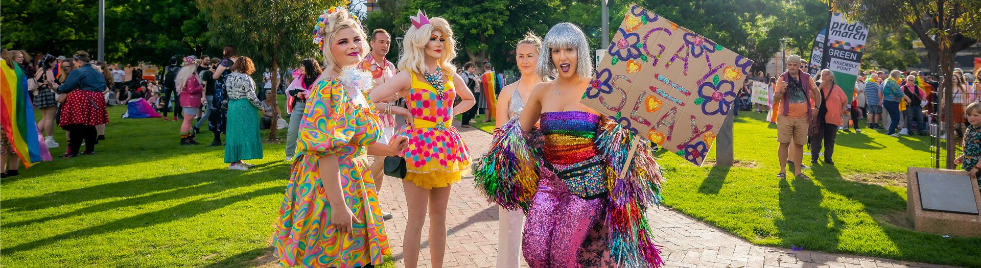 A crowd of people in a park , 4 people in the foreground in colourful outfits, the person on the far right is holding a sign which says 'Gay = Slay'