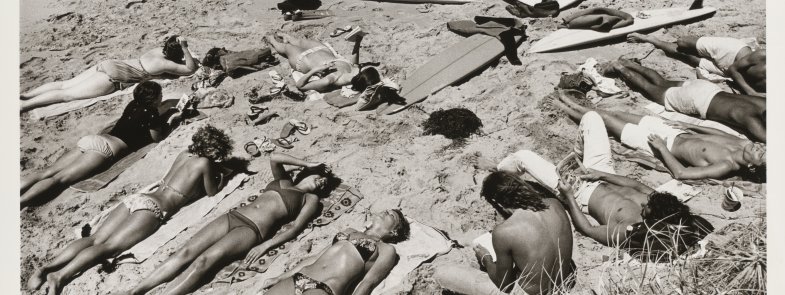 A black and white landscape of a group of sunbathing people lying on a beach with multiple surfboards lying on the ground together. 