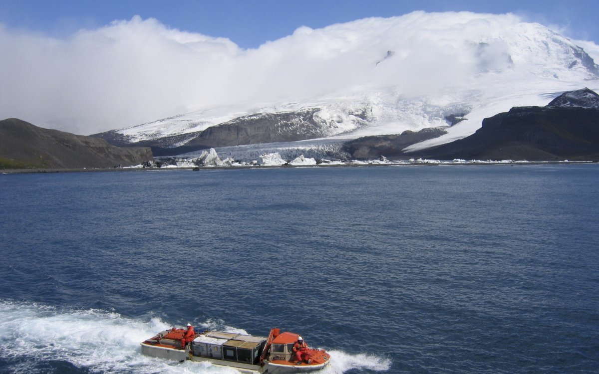 A colour photograph showing snoy mountains with low cloud at their peaks, an expanse of blue water in the foreground with an orange boat on it.