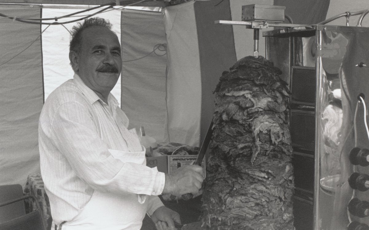 A man with a dark moustache and a white apron is carving meat from a revolving pile to be used for kebabs. He is inside a tent.