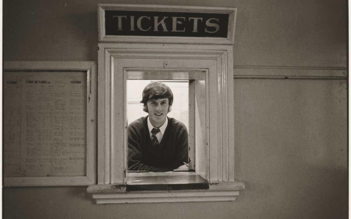 A young man leaning on the ledge of a ticket booth with a program sitting to his left