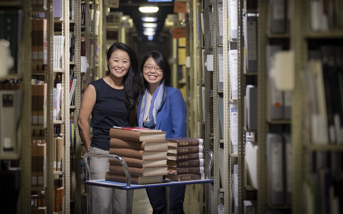 Two women standing behind a trolley with books on it in the Library stacks.