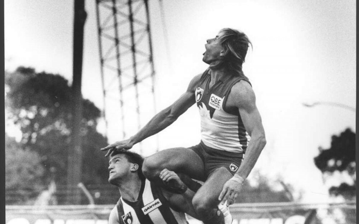 A black and white photo of Warwick Capper jumping high onto the shoulder of another player while looking up in the air.