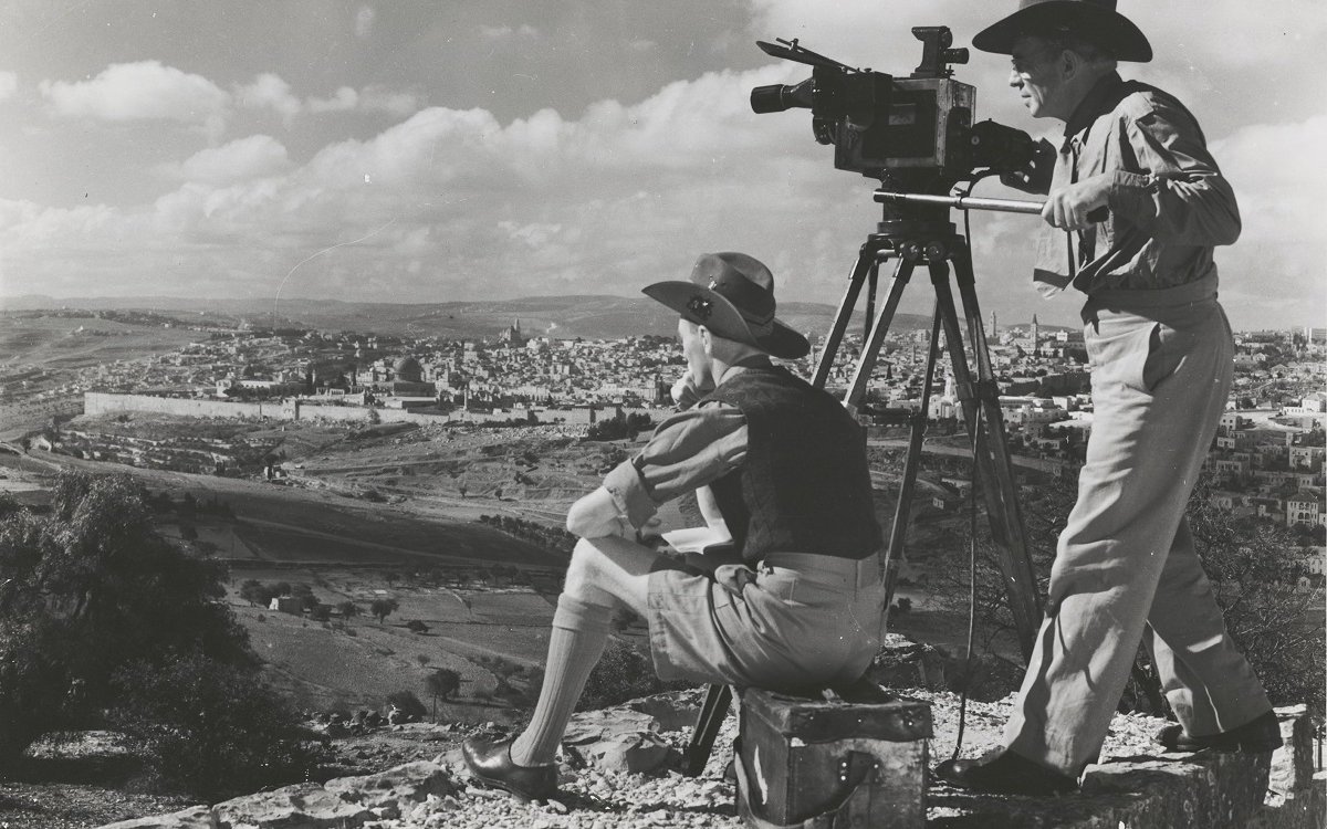 A black and white image of two men on a hilltop overlooking a valley. Both men are wearing hats. One man is sitting on a box. The other is standing up behind an old 1940s style film camera that is on a tripod.