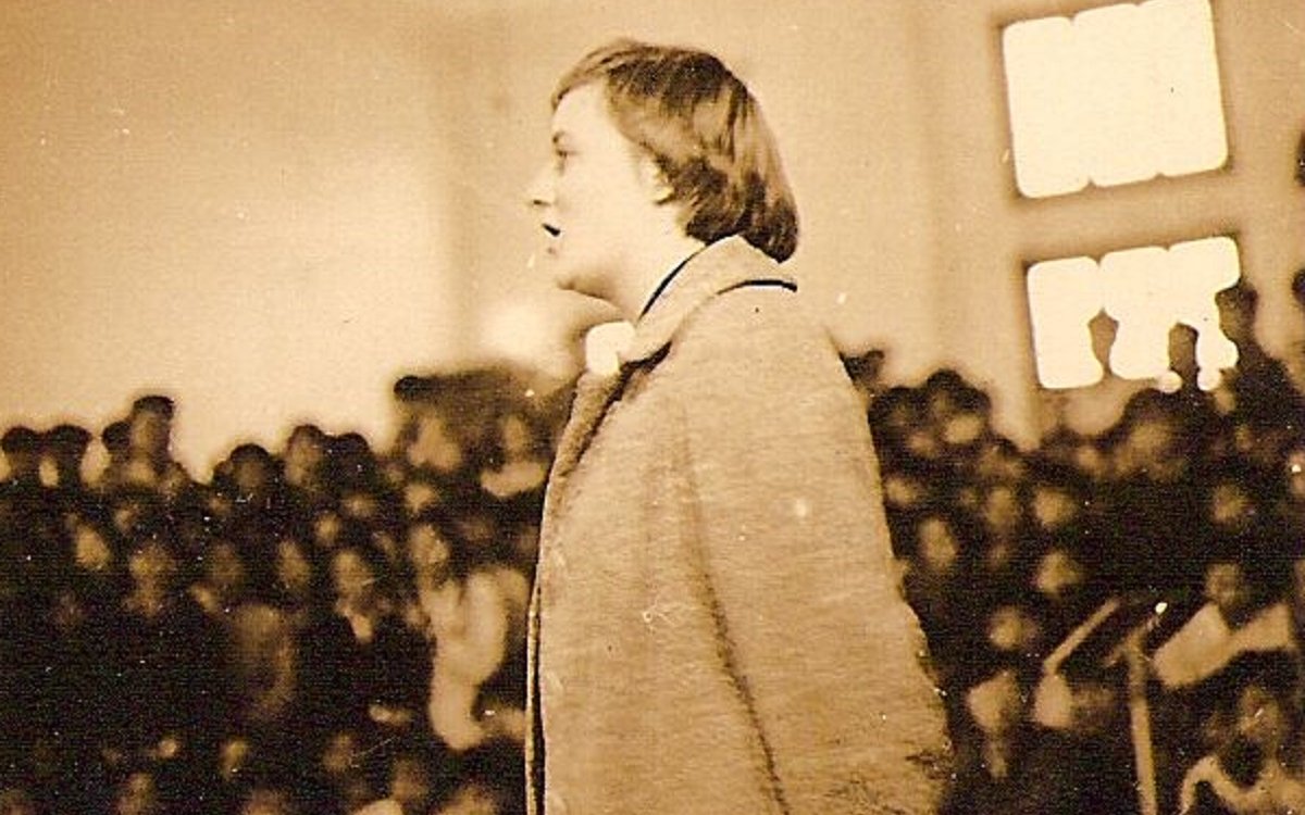 A sepia image of a woman standing up in front of a crowd and speaking.