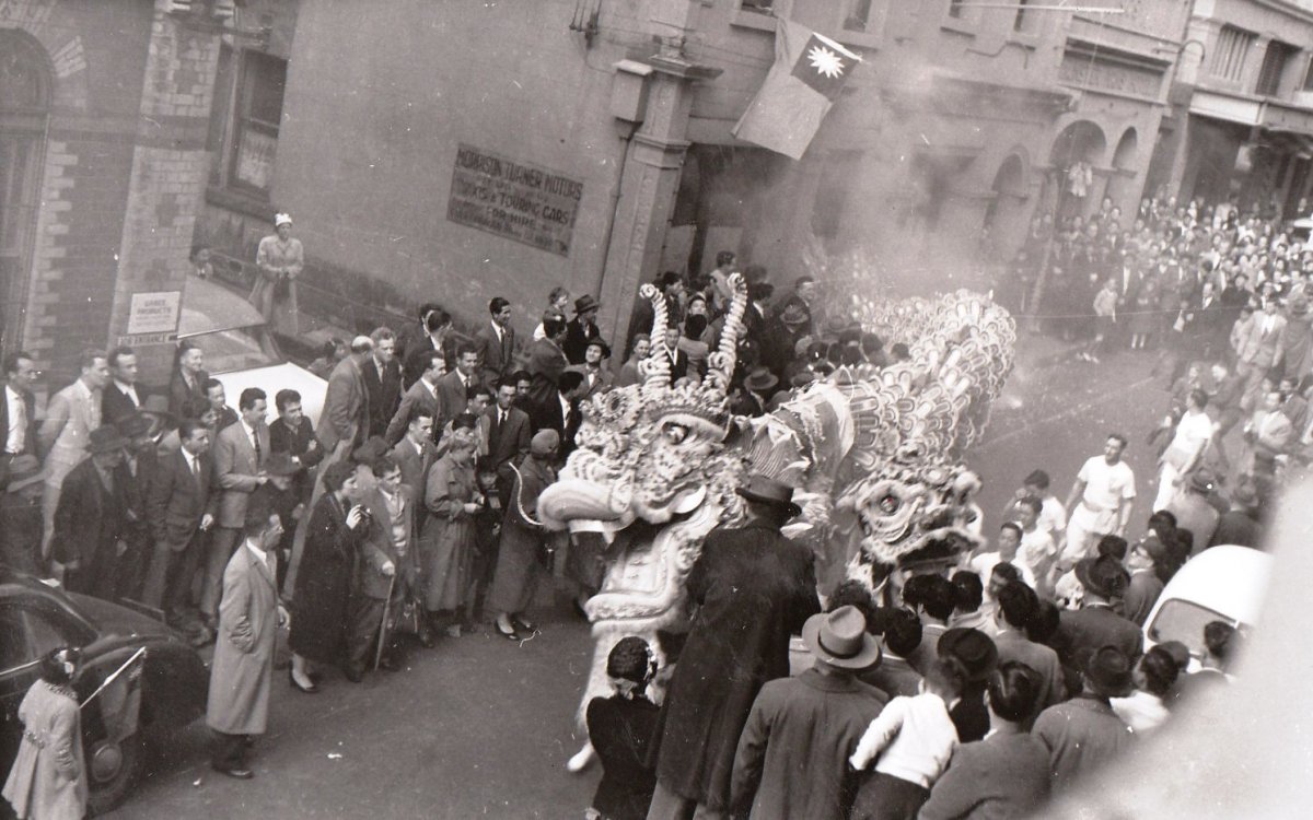 A black and white photograph of a parade down a street. The street is lined with people, and the focus of the parade is a large Chinese dragon.