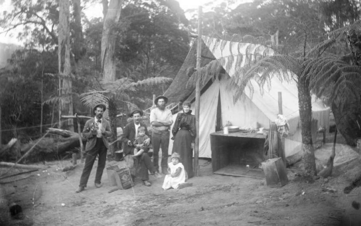 A black and white photograph of a family dressed in late 1800s style fashion. The are standing in a clearing in a forest and are standing in front of a tent.
