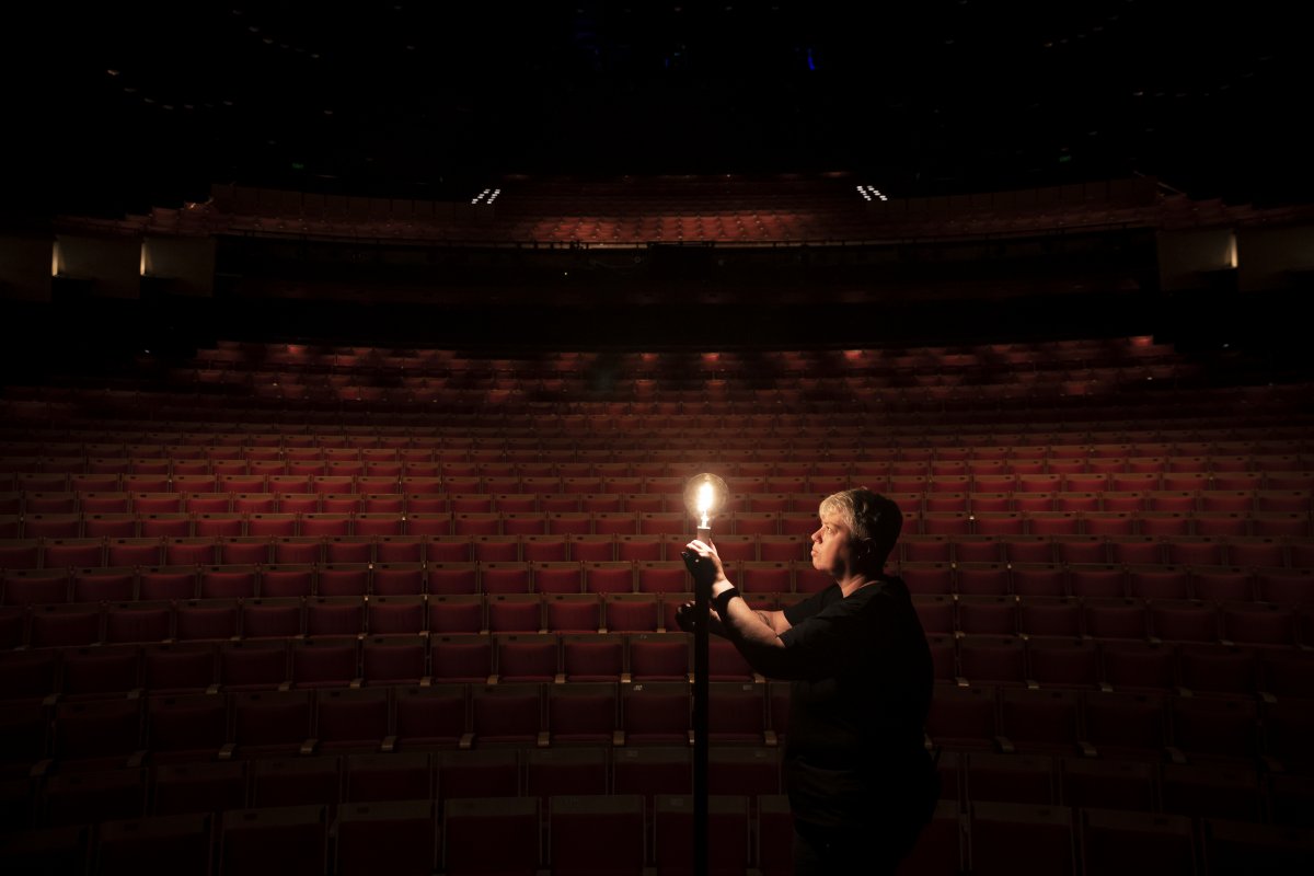 A person stands in an empty darkened theatre, illuminated by a single light bulb.
