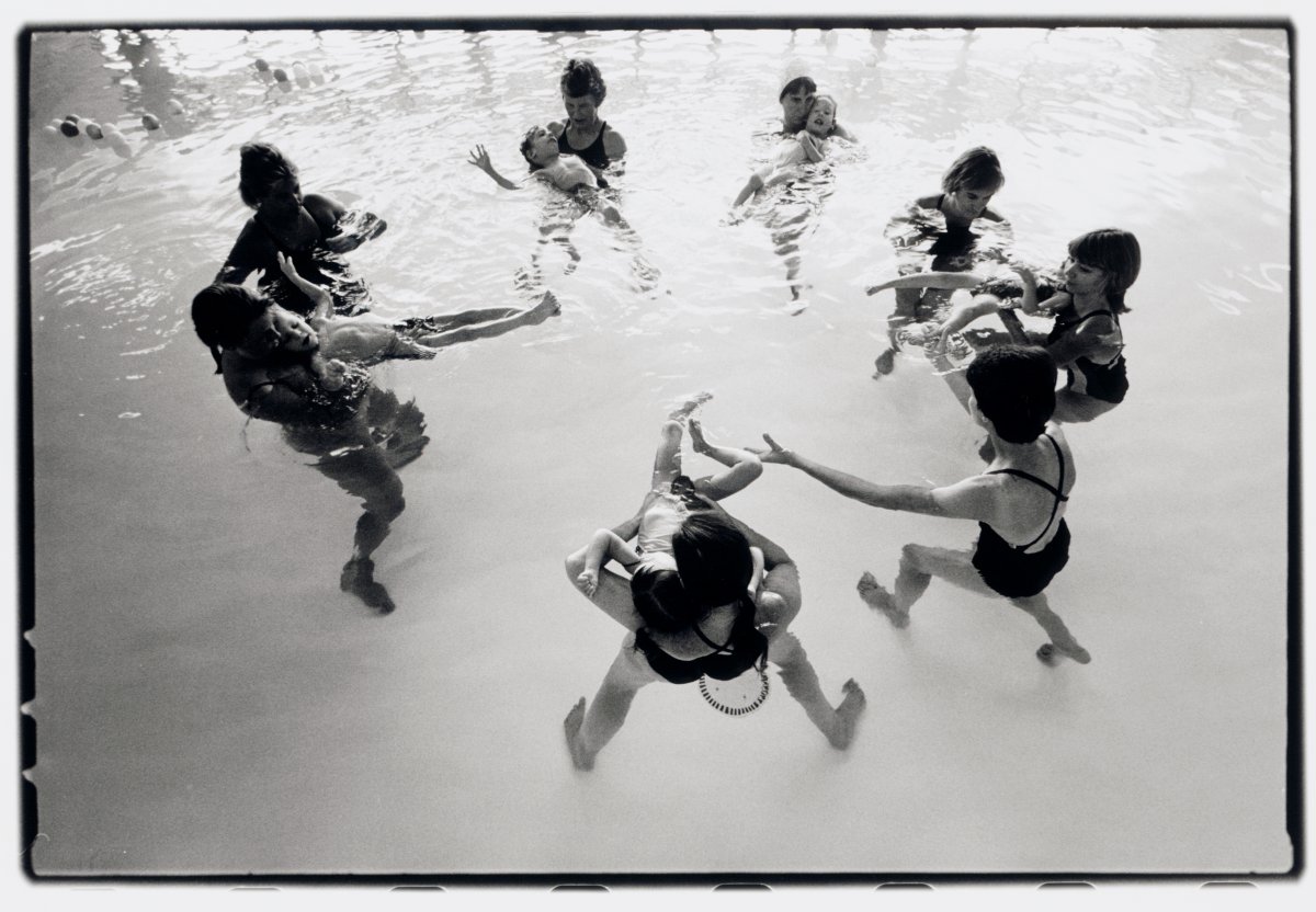 A black and white landscape of women floating in a pool in a circle with toddlers accompanying them. 