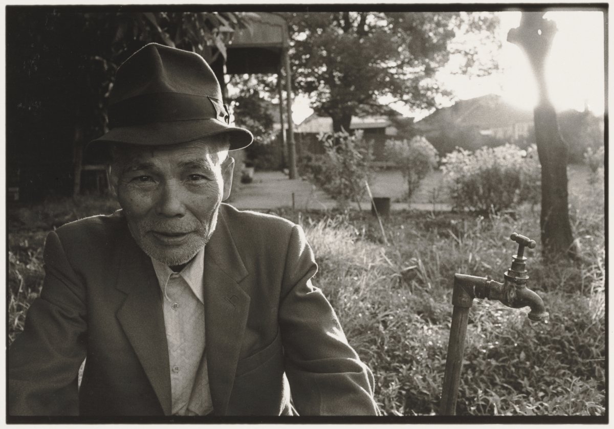 A black and white landscape of an older man wearing a hat and suit jacket sitting outside next to a water tap