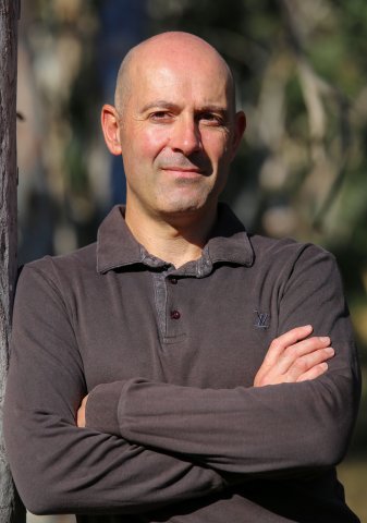 An image of a bald man in a long-sleeved, collared shirt. He has his arms folded across his chest.