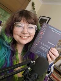 A woman with coloured hair and glasses sitting behind a microphone holding a book up next to her face.