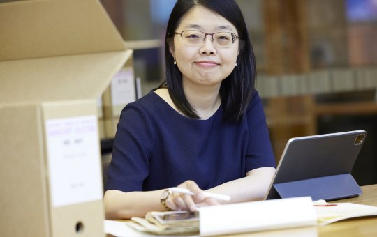 Woman with short dark hair wearing a navy short sleeve top smiling at the camera and sitting at a desk with a laptop, papers and a box