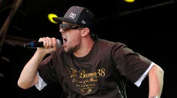 A man sings into a microphone. He is wearing a cap, sunglasses and tshirt.