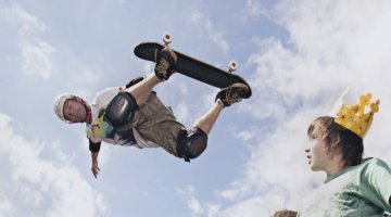 A person on a skateboard is in the air. Two people in the bottom right hand corner are looking up at him. One of them is wearing a cardboard crown. The background is a blue sky with white clouds.