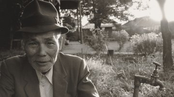 A man on the left staring at the camera sitting outside with an outdoor tap to his right. He wears a hat, a suit jacket and a white dress shirt