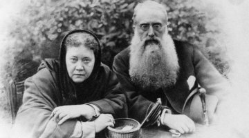 A black and white image of a woman and a man sitting down at a table. There is some kind of shrubbery in the background. The woman is wearing a head scarf covering. The man has a long beard and wears glasses. He is holding a cane and a hat. On the table is an small wicker basket that has been opened, and there is something indistinguishable inside it. At the bottom of the image, under each person, are their signatures.
