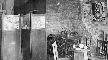 A black and white image of a room with tables and chairs, a wooden room divider, and a modern artwork on the brick wall.