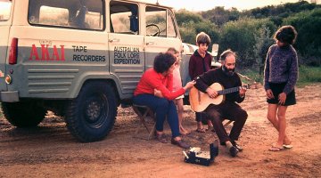 Five people sitting and standing next to a four wheel drive car. One of the people sitting is playing guitar.