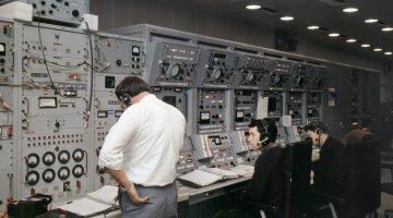 One man standing and two sitting in front of wall of dials, buttons and lights