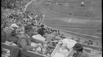 Family of three sitting in the crowded stands of a sports stadium while various events take place on the track and field. The parents seem to be enjoying a warm beverage while looking at their baby who lies in a stroller.