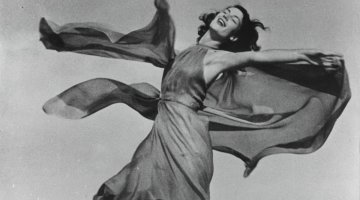 A black and white photograph of a woman in a dress twirling around with her eyes closed.