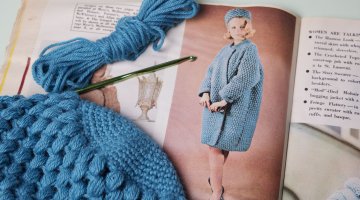 An old magazine showing a vintage crochet pattern. Laying on top of the magazine is a blue crochet item in progress.