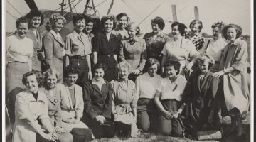 Black and white photo of 21 women of various ages smiling, standing in front of a plane. Below the image is a list of these women's names and where they're from. 