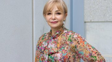 Portrait photograph of a woman with blonde hair, wearing a colourful floral shirt and with one hand on her hip. 