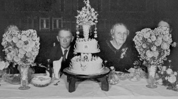 Husband and wife sitting at a table with a large cake and vases of flowers in front of them