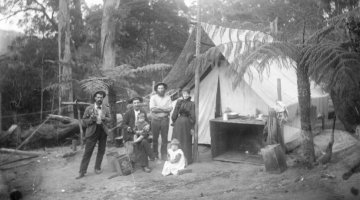 A black and white photograph of a family dressed in late 1800s style fashion. The are standing in a clearing in a forest and are standing in front of a tent.