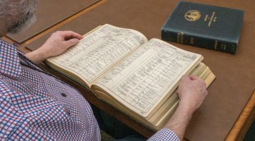 Man reading through a thick, leather-bound book of shipping registers