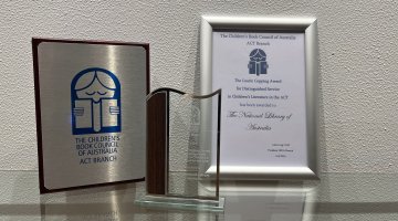 A silver and wooden plaque, glass trophy and framed certificate for the 2024 CBCA Laurie Copping Award given to the National Library