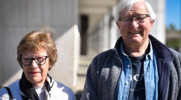 A man and woman, both Library volunteers, standing outside the Library on a sunny day smiling