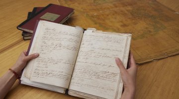 Hand holding an old notebook with handwritten text. In the background is a stack of books and an old map.