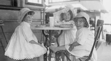 Three young girls in white summer dresses and hats sitting at a cafe table