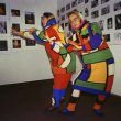 Two people dressed in bright coloured knitted garments pose in front of a wall of photos