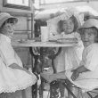 Three young girls in white summer dresses and hats sitting at a cafe table