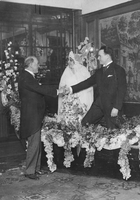 A couple in wedding dress shake the hand of a third man in a suit