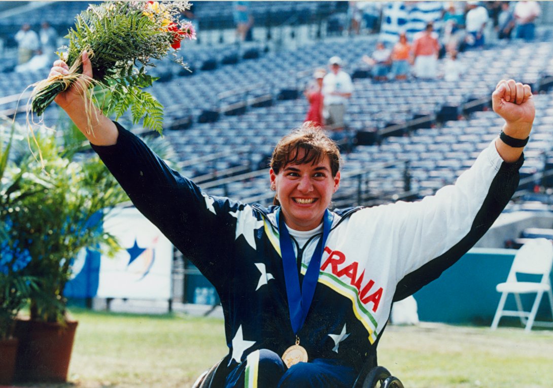 A seated woman faces out of the photograph with her arms in the air has a bunch of flowers in her right hand and a gold medal on a blue ribbon around her neck. Behind her, some grass is visible and stadium seating can be seen behind that. Her jumper has white stars on a navy background and says 'Australia' on it in red capital letters