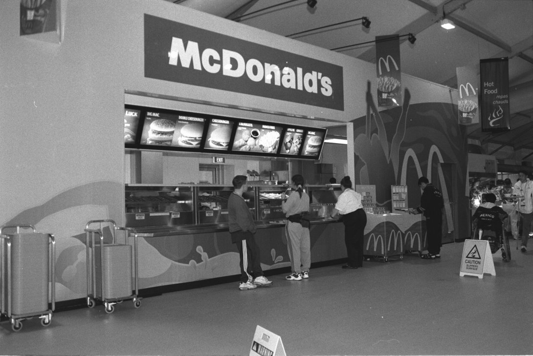 Three people in tracksuits stand in front of a McDonalds counter that seems to be in a bigger room with other food outlet options.