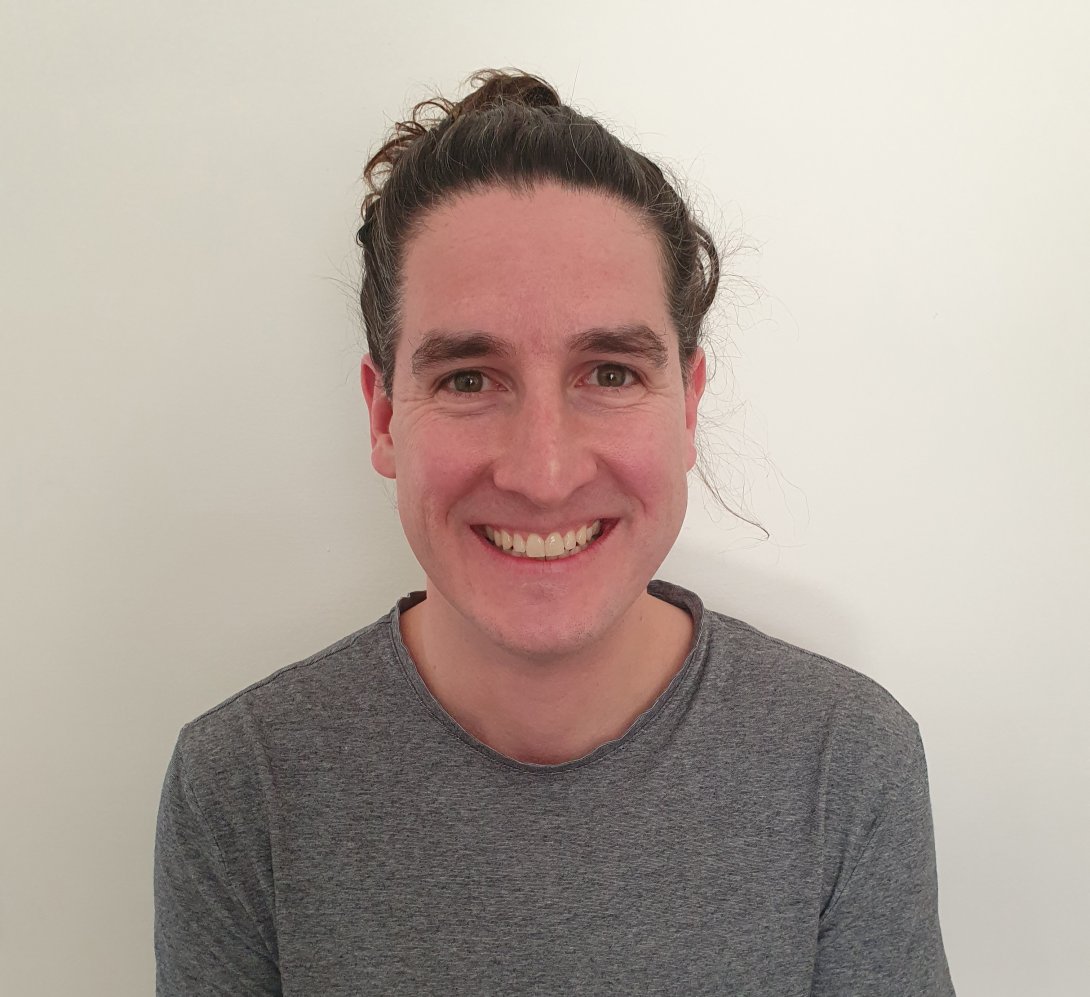 A smiling man with long dark hair tied in a bun looks directly into he camera. He is wearing a grey t-shirt