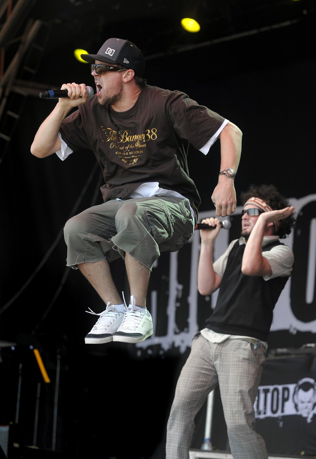 A man in shorts, tshirt, cap and sunglasses is depicted jumping in mid-air, singing into a microphone. Another musician stands behind him.