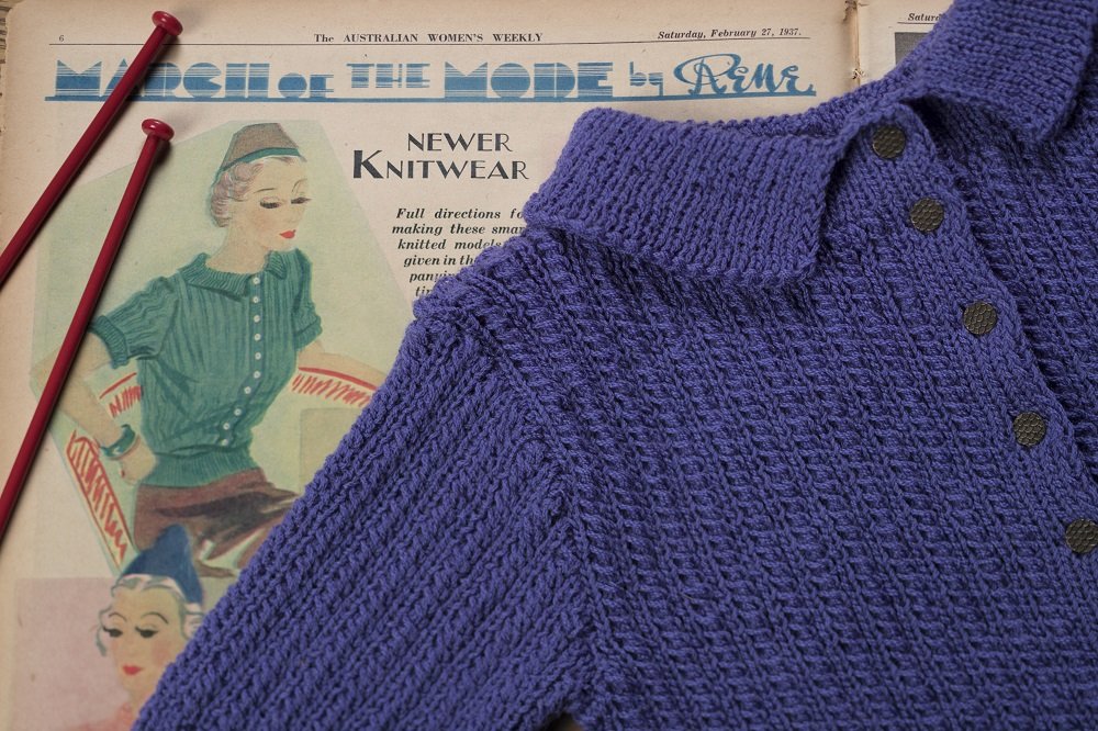 Knitting needles and a knitted blue garment rest on a vintage knitting pattern.