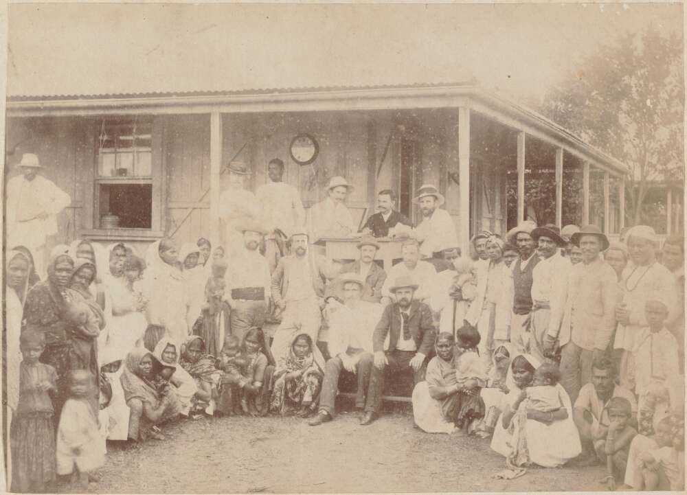 A faded sepia photograph shows a large group of people in front of a house