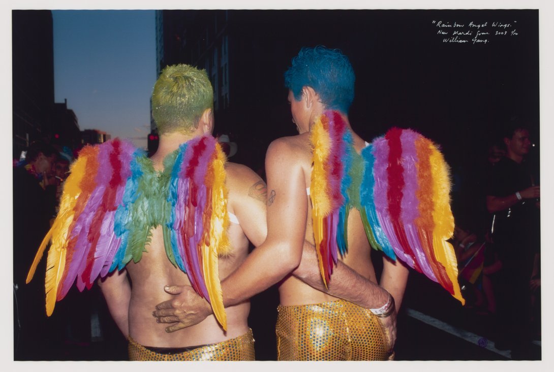 Two men from the waist up with their backs to the camera wearing rainbow wings and no shirts. Their arms are wrapped around each other's waist. 