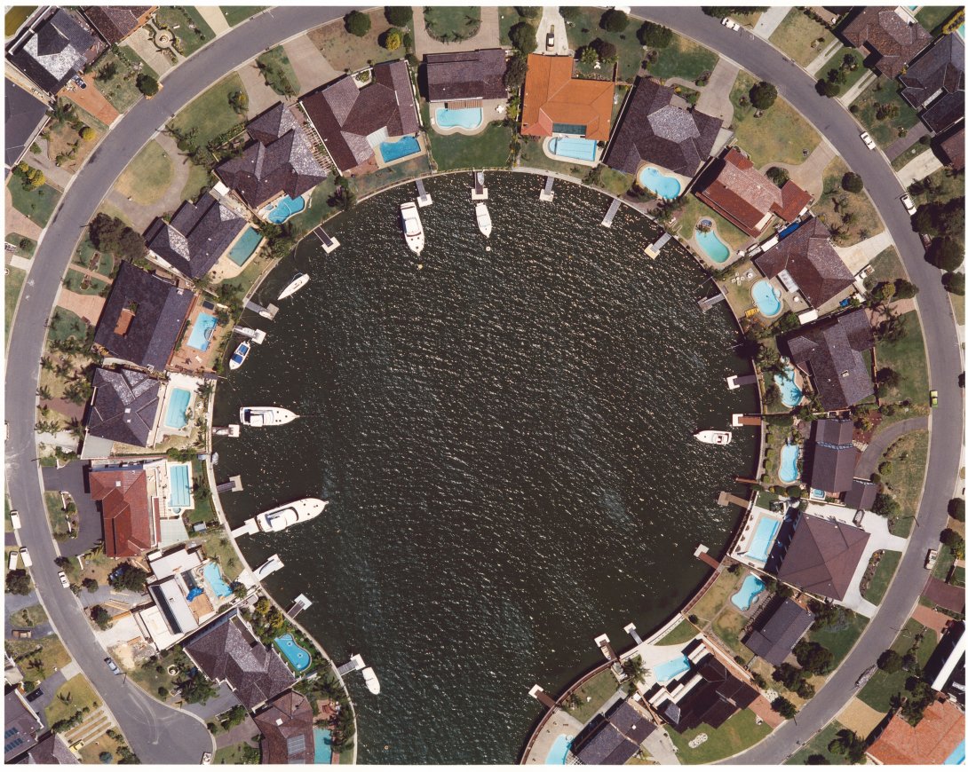 An aerial shot of a suburb. The houses sit in a circle around a round body of water with an opening for the water at the base of the image. there are boats at piers in the water.