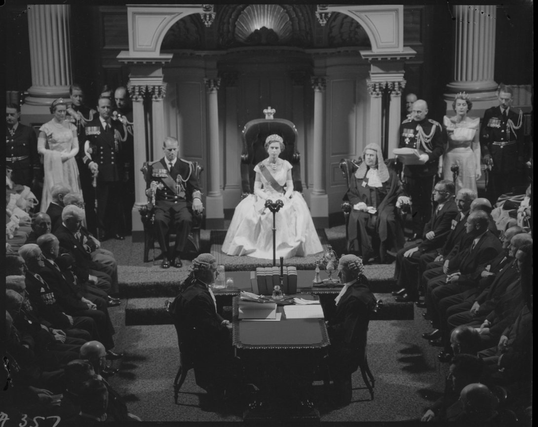 A black and white image of the Queen in full formal attire with tiara sitting in an ornate chair on a dias. There are people sitting on chairs  on either side of the floor below the dais. There are also two people sitting on opposite sides of a table on that is directly in front of the dais.